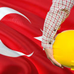 Turkish Engineer is holding yellow safety helmet with waving Turkey flag background. Construction and building concept.
